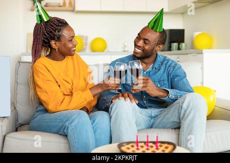 Liebende schwarze Ehepartner, die ein Glas Wein trinken, zu Hause Geburtstag feiern, auf dem Sofa sitzen und einander anlächeln Stockfoto