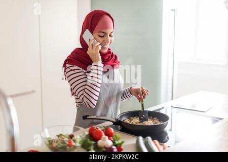 Fröhliche muslimische Hausfrau kocht Abendessen, telefoniert Stockfoto