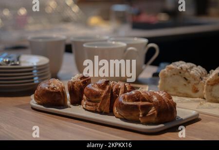 Konzept einer Kaffeepause im Innenbereich mit Zimtbrötchen. Kaffee- und Teetassen und Kuchenscheiben im Hintergrund. Foto wurde in Schweden aufgenommen. Stockfoto