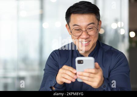 Nahaufnahme eines glücklichen und lächelnden Asiaten, der am Telefon fröhliche Nachrichten liest, ein Geschäftsmann in Hemd und Brille, der lächelt und jubelt, ein Mann arbeitet im Büro neben dem Fenster. Stockfoto