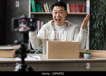 Aufgeregter junger, männlicher asiatischer Blogger, der Geschenkboxen von Unternehmen auspackt und Livestreaming macht. Attraktiver Mann, der eine Smartphone-Kamera zum Erstellen von Inhalten verwendet. Stockfoto