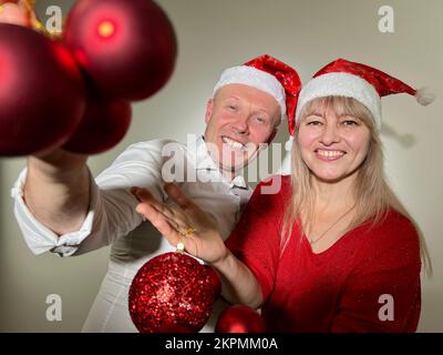 Partyeinladung Neujahr und Weihnachtsmann und -Frau Erwachsene tanzen in den Händen mit Spielzeug, haben Weihnachtshüte auf dem Kopf und rote Bälle Ded Moroz Weihnachtsmann Schneemädchen Stockfoto
