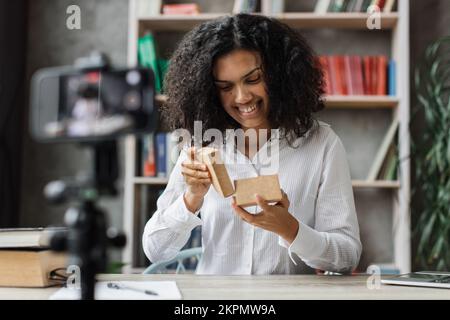 Fröhliche Bloggerin, die Geschenkboxen von Unternehmen auspackt und Live-Streaming macht. Multinationale Frau, die eine Smartphone-Kamera zum Erstellen von Inhalten verwendet. Stockfoto