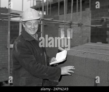 1980er, auf einer Baustelle für Wohnungen, ein Vorarbeiter in einer Eselsjacke und einem Schutzhelm mit einem Notebook, während er einen Stapel leichter Betonbausteine prüft, die beim Bau neuer Wohnungen in England, Großbritannien, verwendet wurden. Stockfoto