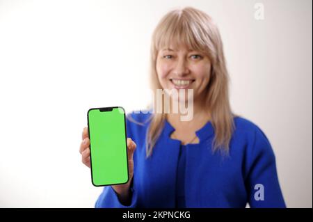 Eine Erwachsene Frau im Alter von 45-50 mit blondem Haar in einem blauen Kleid steht mit einem Handy Grüner Bildschirm Chroma Key, sie lächelt und wirbt für jede Nachfrage nach Produkten Stockfoto