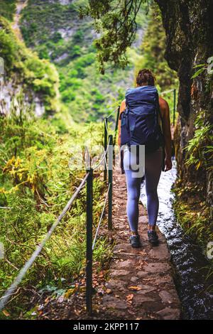 Beschreibung: Sportliche Frau mit Rucksack geht durch abenteuerliche Dschungelwege entlang eines grünen, überwucherten Kanals. Levada von Caldeirão Verde, Insel Madeira Stockfoto