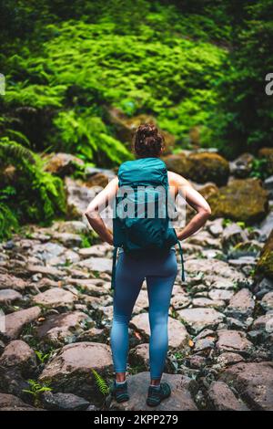 Beschreibung: Sportliche Frau mit Rucksack geht durch abenteuerliche Dschungelwege entlang des felsigen Flussbetts. Levada von Caldeirão Verde, Insel Madeira, Hafen Stockfoto