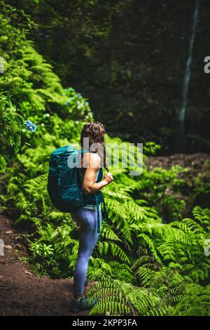 Beschreibung: Sportliche Frau mit Rucksack bewundert einen beeindruckenden Wasserfall im Dschungel mit wunderschönen Blumen und Farnen. Levada Caldeirão Verde, Stockfoto