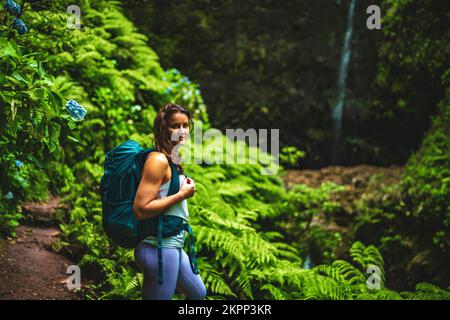 Beschreibung: Sportliche Frau bewundert die Natur an einem beeindruckenden Wasserfall im Dschungel mit wunderschönen Blumen und Farnen. Levada von Caldeirão Verde, gemacht Stockfoto