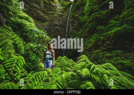 Beschreibung: Wunderschöne sportliche Frau mit Rucksackwanderungen entlang der Blumenstrasse und dem farny Trail am herrlichen Dschungelwasserfall. Levada Caldeirão Verde, Madei Stockfoto