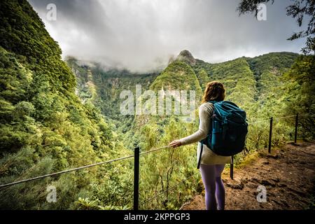 Beschreibung: Wunderschöne Frau mit Rucksack bewundert die Aussicht vom abenteuerlichen Dschungelweg entlang des Kanals. Levada Caldeirão Verde, Madeira Stockfoto