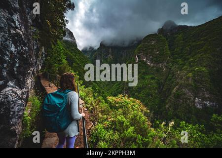 Beschreibung: Wunderschöne Frau mit Rucksack bewundert die Aussicht vom abenteuerlichen Dschungelweg entlang des Kanals. Levada Caldeirão Verde, Madeira Stockfoto