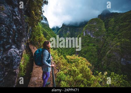 Beschreibung: Wunderschöne Frau mit Rucksack bewundert die Aussicht vom abenteuerlichen Dschungelweg entlang des Kanals. Levada Caldeirão Verde, Madeira Stockfoto