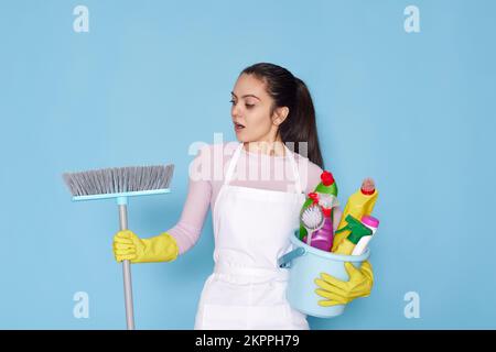 Frau in Handschuhen und Reinigungsschürze mit Eimer mit Waschmitteln und Besen Stockfoto