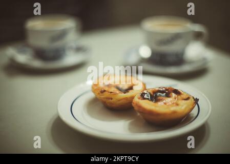 Nahaufnahme von zwei traditionellen portugiesischen Vanilletörtchen und Tassen Kaffee auf einem Tisch Stockfoto