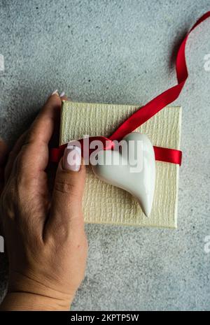 Frauenhand mit einer weißen Geschenkbox, die mit einem roten Band verbunden und mit einem keramischen Herzschmuck verziert ist Stockfoto