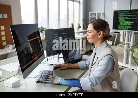 Positiver Softwareentwickler mit bionischem Arm, der Code auf dem Computerbildschirm auf Fehler prüft Stockfoto