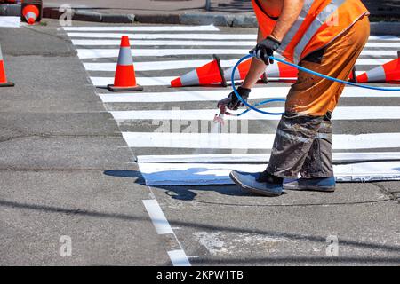 Ein Straßenarbeiter bringt weiße Straßenmarkierungen auf einem Fußgängerüberweg mit einer Luftbürste und einer Holzschablone an, die von Verkehrskegeln umgeben sind. Stockfoto