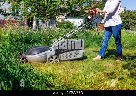 Eine Gärtnerin schneidet im Garten hohes grünes Gras. Ein Gärtner mäht hohes Gras mit einem elektrischen Rasenmäher im Garten. Stockfoto