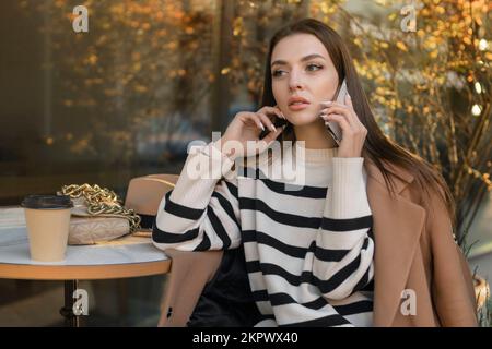 Eine brünette, wunderschöne, stilvolle, weiße Frau in lässiger Kleidung auf einem Spaziergang entlang der Straße einer europäischen Stadt sitzt in einem Café und spricht am Telefon Stockfoto