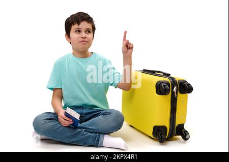 Ein niedlicher Junge in blauen Jeans und lässigem T-Shirt, der neben dem gelben Koffer sitzt und eine Bordkarte in der Hand hat Stockfoto