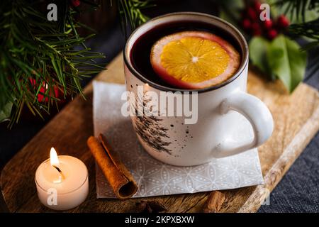 Mulled wine with cinnamon and orange served in festive Christmas decoration on wooden board. Party and New Year atmosphere. Christmas tree in backgrou Stockfoto