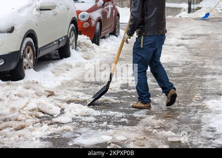 Ein Mann reinigt Schnee und Eis mit einer Schaufel von einem Asphaltweg in der Nähe geparkter Autos im Hof des Hauses. Stockfoto