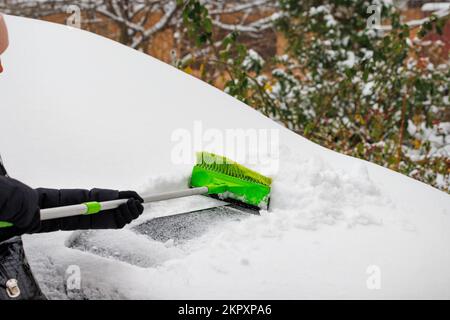 Eine Frau bürstet Schnee von der Motorhaube eines Autos. Ein Mann reinigt nach einem Schneesturm im Winter frischen Schnee von einem Auto. Stockfoto