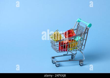 Nahaufnahme eines kleinen Einkaufswagens voller farbenfroher Weihnachtsgeschenke auf blauem Hintergrund. Speicherplatz kopieren. Stockfoto