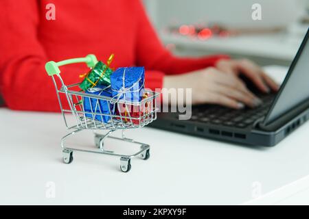 Einkaufswagen mit bunten Geschenken auf dem Tisch. Frau, die ein Notebook im Hintergrund benutzt. Während der Feiertage online einkaufen. Stockfoto