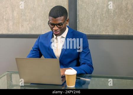 Lächelnder afrikanischer Geschäftsmann in Anzug, Brille, die vom Büro aus an einem Laptop arbeitet. Schwarzer Mann, der Computer benutzt, am Schreibtisch sitzt und ein Webinar ansieht Stockfoto