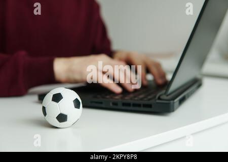 Junger Mann liest Fußballnachrichten oder schreibt Fußballartikel auf einem Laptop. Fußball auf dem Tisch. Wetten, Glücksspiel. Stockfoto