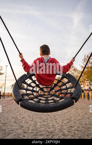 Kleiner Junge, der im Park spielt oder auf einer runden Spinnennest-Schaukel schwingt. Freizeitaktivitäten für Kinder im Freien. Stockfoto