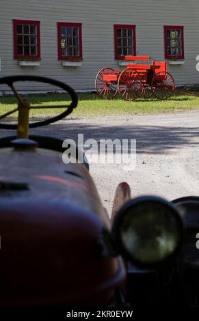 Ein alter Farmtraktor und ein roter antiker Wagen neben einer alten weißen Scheune. Stockfoto