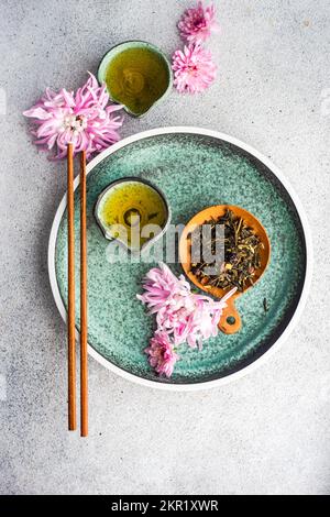 Blick von oben auf zwei Tassen grünen Tee und Teeblätter auf einem Teller mit pinkfarbenen Chrysanthemen Stockfoto