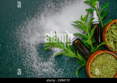 Süße Pflanze. Stevia rebaudiana. Trockener Stevia, Pulver und flüssiger Stevia-Extrakt in einer Glasflasche auf dunkelgrünem Hintergrund. Natürlicher Zuckerersatz Stockfoto