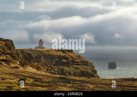 Blick auf den Atlantischen Ozean von Dyrhólaey, mit dem Leuchtturm von Dyrhólaeyjarviti in der Ferne, unter einem dramatischen bewölkten Himmel, Island Stockfoto