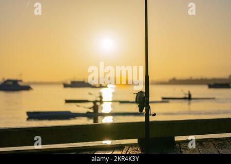 Angelrute und -Rolle stehen bei Sonnenaufgang auf dem Anlegesteg und hoffen auf einen Fang an der Küste von Tauranga am Anlegesteg. Wie unscharfe Boote und Kajaks B bilden Stockfoto