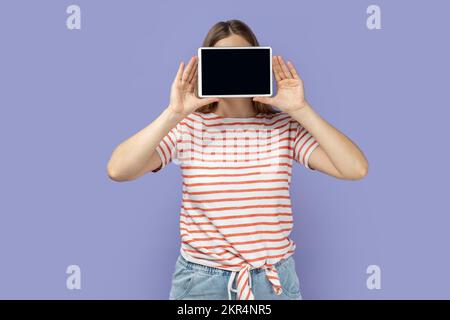 Porträt einer anonymen, unbekannten blonden Frau, die gestreiftes T-Shirt trägt, das Gesicht mit einem Tablet bedeckt, mit leerem Display, Kopierraum für Werbetext. Studio-Aufnahme im Innenbereich isoliert auf lila Hintergrund. Stockfoto