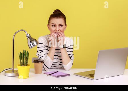 Ängstliche, junge Büroleiterin, die sich die Nägel kaut, sitzt am Arbeitsplatz und sieht mit nervöser Sorge in die Kamera. Studioaufnahmen im Innenbereich isoliert auf gelbem Hintergrund. Stockfoto