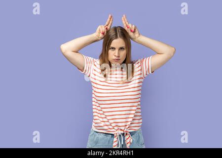 Porträt einer wütenden, herrisch blonden Frau, die gestreiftes T-Shirt trägt, im Stehen Hörner zeigt, wütend und aggressiv ist, in die Kamera schaut. Studio-Aufnahme im Innenbereich isoliert auf lila Hintergrund. Stockfoto