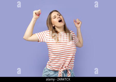 Porträt einer müden blonden Frau, die ein gestreiftes T-Shirt trägt, mit geschlossenen Augen gähnt, Arme streckt, sich erschöpft fühlt, Ruhe braucht. Studio-Aufnahme im Innenbereich isoliert auf lila Hintergrund. Stockfoto