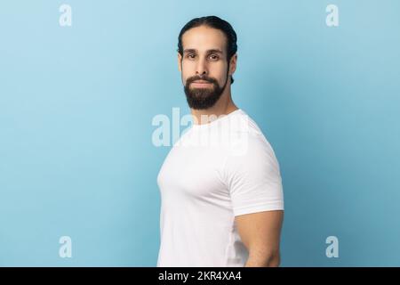 Seitliches Porträt eines bärtigen, gutaussehenden Mannes, der ein weißes T-Shirt trägt, in die Kamera schaut, mit ernstem Gesicht, streng und rechthaberisch ist. Innenstudio, isoliert auf blauem Hintergrund. Stockfoto