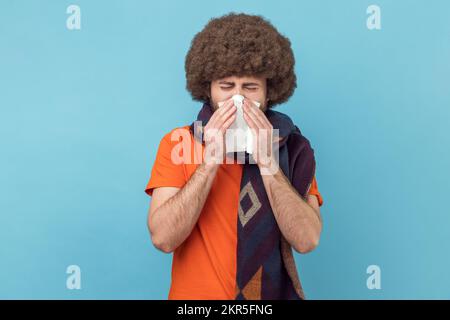 Porträt eines ungesunden Mannes mit Afro-Haarschnitt, der ein orangefarbenes T-Shirt trägt, das Gewebe auf Mund und Nase hält, in einen warmen Schal gewickelt wird. Innenstudio, isoliert auf blauem Hintergrund. Stockfoto