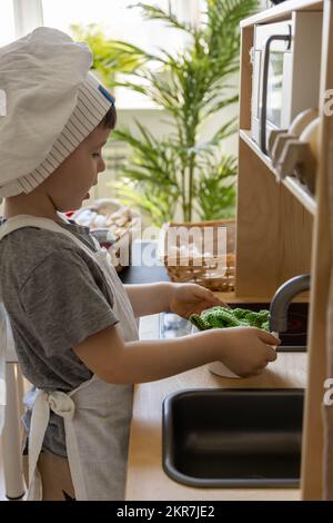 Niedlicher kleiner Junge mit Kochmütze und Schürze, der in der kindischen Küche beim Kochen von Speisen spielt Stockfoto