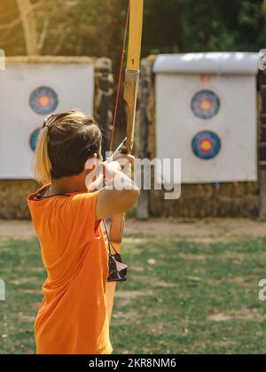 Rückansicht der asiatischen Jungen tragen Gesichtsmaske zielt Bogenschießen Bogen und Pfeil zu bunten Ziel im Schießstand während des Trainings und des Wettbewerbs. Übung und Co Stockfoto