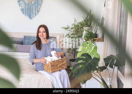 Eine glückliche junge Frau im warmen Bademantel hält einen Stapel gerollter Bio-Baumwollhandtücher in einem weißen Korb Stockfoto