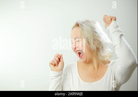 Einladung Werbung Party Ort für Sie Erwachsene Frau tanzt sie lächelt mit offenem Mund und erhobenen Armen viele positive Emotionen gewinnen T-Shirt Hintergrund und weiße Haare Stockfoto