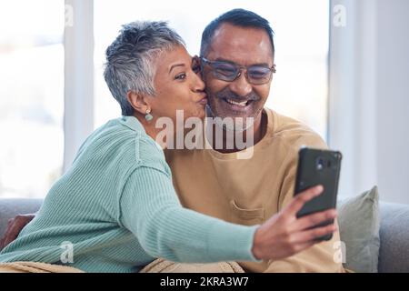 Telefon, Selfie und Kuss mit einem älteren Paar, das für ein Foto posiert, im Wohnzimmer ihres gemeinsamen Zuhauses. Smartphone, Küssen und soziale Medien mit Stockfoto