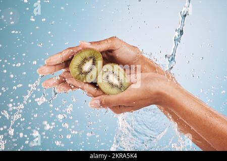 Wasserspritzer, Frauenhände und Kiwi im Studio auf blauem Hintergrund. Reinigung, Hygiene und weibliche Modellwäsche Früchte für gesunde Ernährung, Ernährung Stockfoto